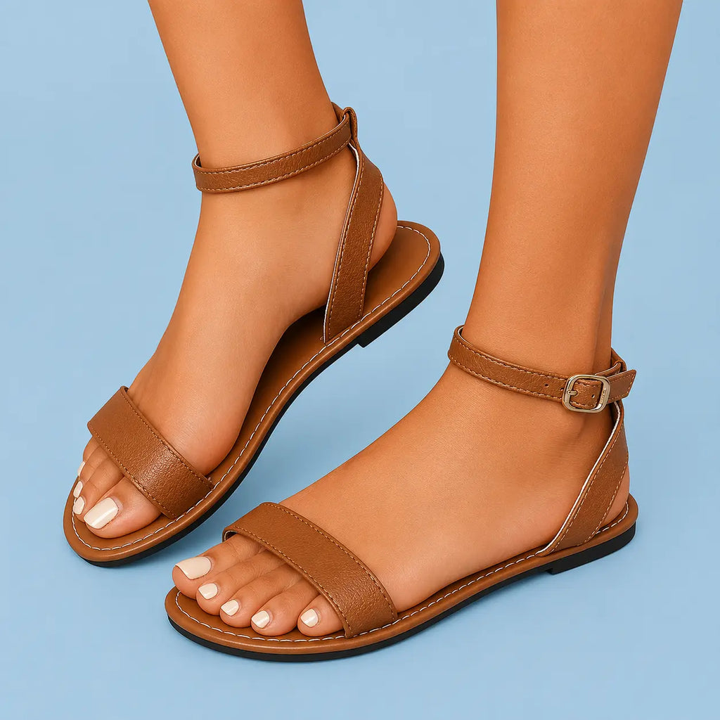 A pair of women's flat strappy sandals in a warm tan-brown color, shown on a model's feet against a solid light blue background.