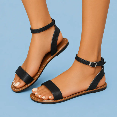 A pair of black flat sandals with ankle straps and a single front strap, shown on a model's feet against a light blue background.